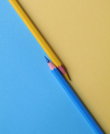 A yellow colored pencil and a blue colored pencil pointed toward each other on blue and yellow background